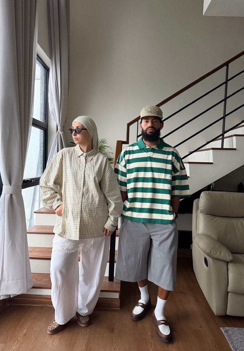 Un homme en polo rayé vert et short gris se tient à côté d'une femme en chemise beige à carreaux, pantalon blanc et foulard sur la tête près des escaliers à l'intérieur.