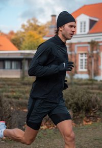 Fekete futódzseki cipzárral, illeszkedő fekete sort és kesztyűk. Futó fekete sapkát visel, és világos színű sportcipő van rajta. Őszi háttér.