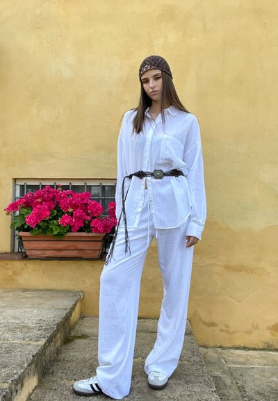 Camisa de lino blanca y pantalones de pierna ancha combinados con un cinturón trenzado marrón y un pañuelo para la cabeza. Zapatos grises con rayas negras. Flores rosas en una maceta.