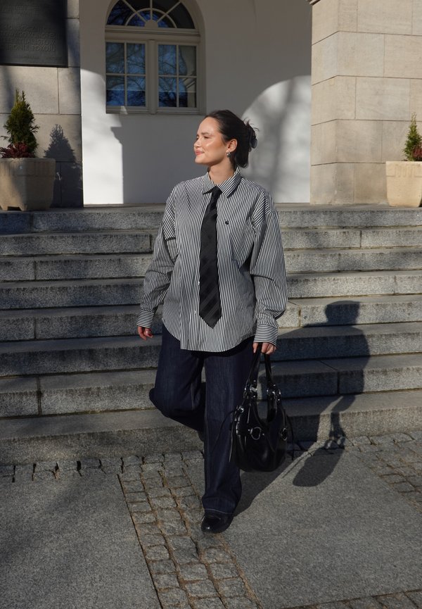 Kvinne i stripet skjorte, svart slips og mørke bukser går ned steintrapper mens hun holder en svart håndveske i sollys.