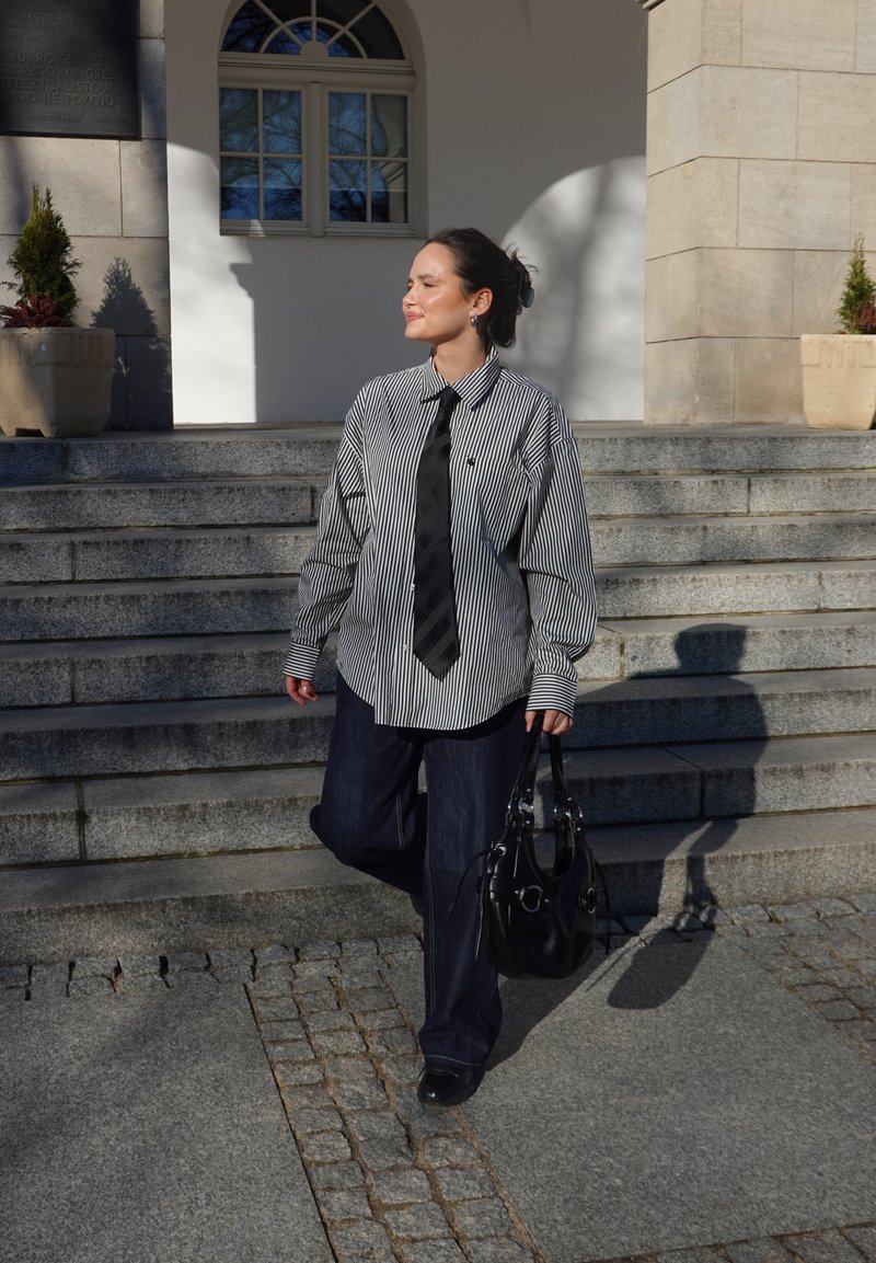 Vrouw in gestreept shirt, zwarte stropdas en donkere broek loopt in het zonlicht een stenen trap af terwijl ze een zwarte handtas vasthoudt.