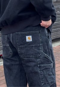 Pantalon cargo en denim noir avec plusieurs poches, coutures renforcées et un patch logo de marque bien visible sur la poche arrière.