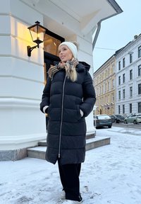 Svart pufferjakke med hette, glidelås og hvite fleece-fôrede lommer. Kombinert med en hvit strikkelue og et beige rutete skjerf. Snøfylt bakgrunn.