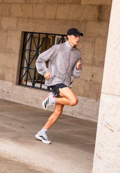 Corredor con una chaqueta impermeable gris claro, pantalones cortos negros y zapatillas deportivas blancas con acentos rojos, corriendo sobre una superficie de piedra.