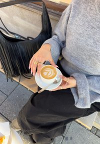 Fehér csészében lévő latte, leveleminta a habon, egy aranygyűrűkkel díszített kéz által egy tányéron tartva. Háttérben fekete himbálós kézitáska.