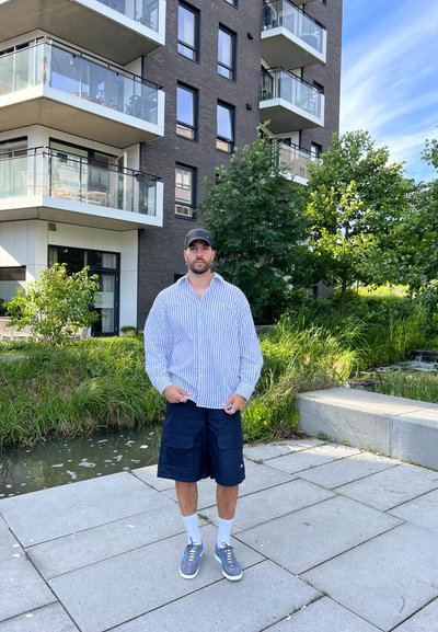 Camisa de rayas azul y blanca de gran tamaño, pantalones cortos cargo azul marino, calcetines blancos, zapatillas grises; de pie en un área pavimentada cerca de un cuerpo de agua.