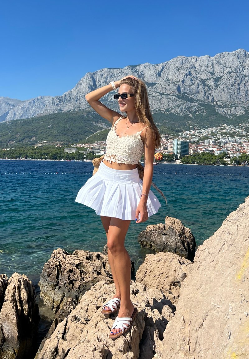 Άσπρη πλισέ φούστα με ένα εφαρμοστό, υφασμάτινο άσπρο μπλουζάκι. Στεκόμενη σε βράχια δίπλα στο νερό, φορώντας γυαλιά ηλίου και άσπρα σανδάλια.