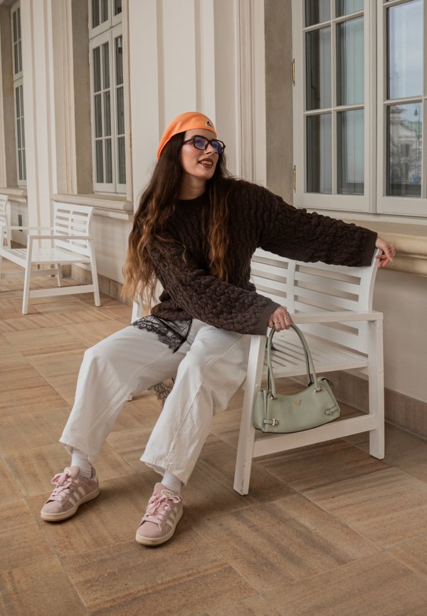 Tânără purtând un beret portocaliu și un pulover maro, stă pe o bancă albă ținând o geantă verde mentă într-un hol cu ferestre mari.