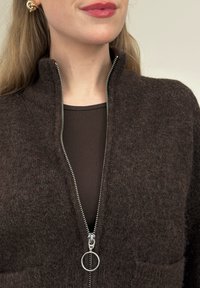 Femme portant une veste zippée en tissu pelucheux marron foncé avec une fermeture éclair argentée et un tire-zip circulaire, associée à un haut côtelé marron foncé et des boucles d'oreilles nœud dorées.