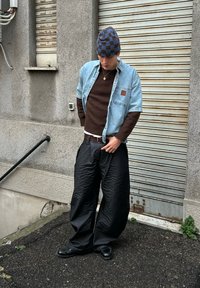 Chemise en jean claire boutonnée, pull marron, pantalon large noir texturé, bonnet à carreaux bleu et noir, accessoirisé avec un collier en or.