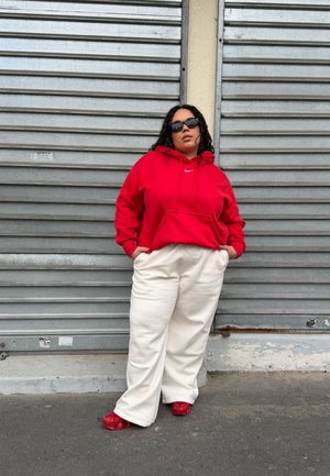 Persona con sudadera con capucha roja, pantalones blancos, zapatos rojos y gafas de sol negras, de pie con las manos en los bolsillos frente a una persiana metálica cerrada.