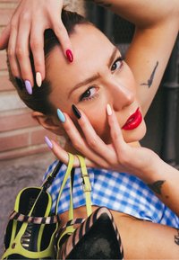 Gros plan sur une personne tenant un sac à main vert, portant un vernis à ongles pastel et vif, avec un haut à carreaux bleu et un tatouage visible.