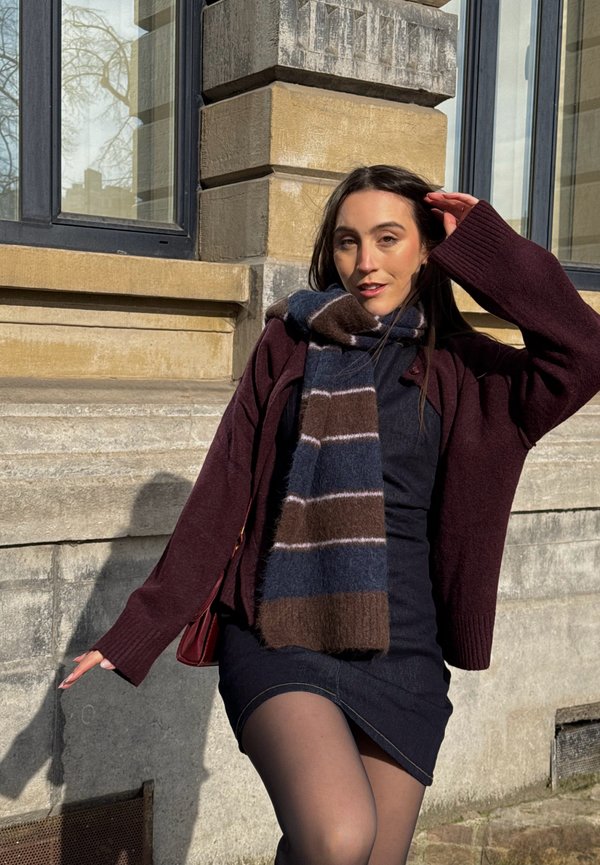 Femme portant une robe sombre, un cardigan bordeaux et une écharpe rayée, posant en extérieur contre un mur en pierre et une fenêtre baignés de soleil.