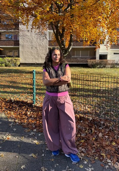 Persona con chaleco sin mangas de piel sintética oscura sobre una camisa rosa, pantalones anchos de color rosa claro y zapatillas azules, con una bolsa verde.