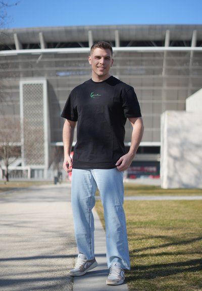 Joven de pie al aire libre con una camiseta negra informal, jeans azul claro y zapatillas deportivas, con un edificio moderno al fondo.