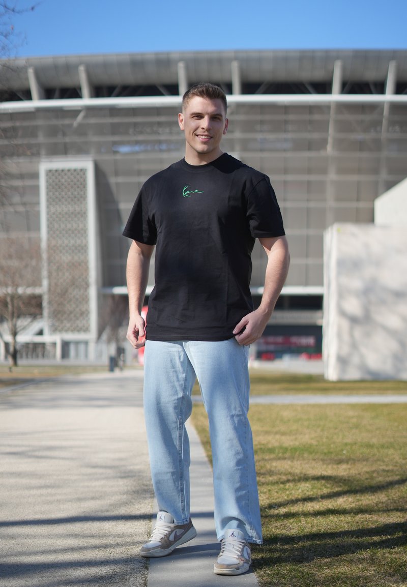 Jeune homme debout en extérieur, portant un t-shirt noir décontracté, un jean bleu clair et des baskets, avec un bâtiment moderne en arrière-plan.