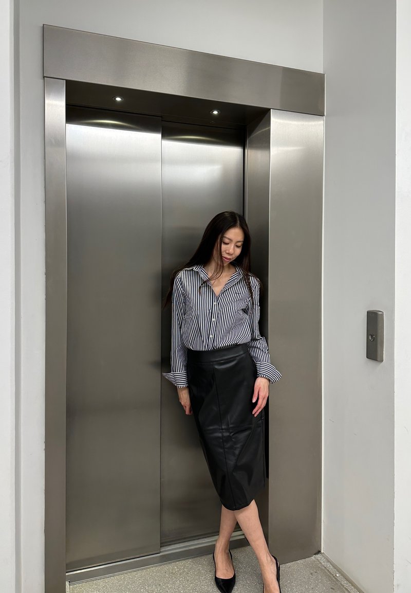 Prugasta srajca s gumbi, črna usnjena svilenka in črne pete. Stoji ob kovinskih dvigalih z vtkanimi lučmi. Gladke teksture.