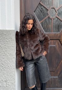 Femme aux cheveux bouclés portant une veste en fourrure brune, un bermuda en cuir noir et des bottes noires, adossée à un mur texturé près d'une porte en bois.