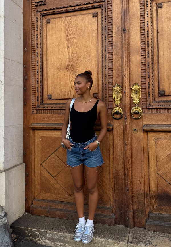 Donna in top nero e pantaloni di jeans corti sta in piedi con le mani nelle tasche accanto a una grande porta in legno ornata con maniglie dorate.