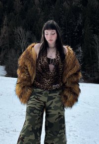 Jeune femme vêtue d'un haut à imprimé léopard, d'une veste en fourrure marron et d'un pantalon camouflage, debout dans un champ enneigé avec une forêt en arrière-plan.