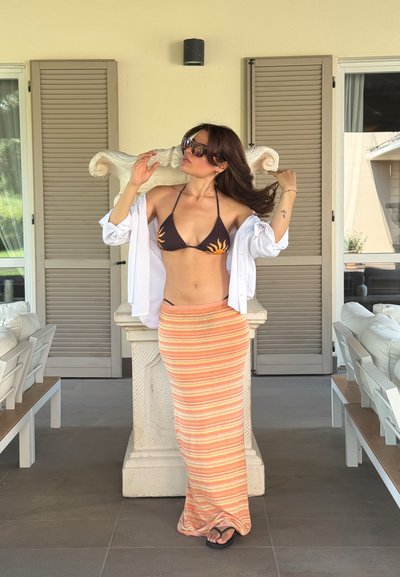 Bikini marrón con diseño de sol naranja, camisa blanca abierta y falda maxi a rayas naranja. Chanclas negras y gafas de sol de gran tamaño.