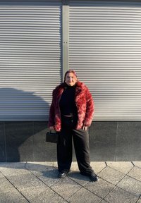 Veste en fausse fourrure rouge, col roulé noir, larges pantalons noirs et sac à main noir. Textures lisses ; motif en peluche ébouriffée sur la veste.