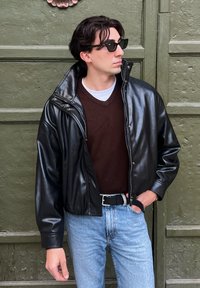 Jeune homme portant une veste en cuir noire, un pull marron, un jean bleu clair et des lunettes de soleil, debout devant une porte verte texturée.