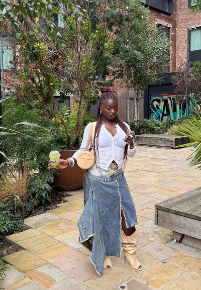 Joven con cabello trenzado que lleva una blusa blanca, una falda de mezclilla y botas beige, sosteniendo una bebida y usando un smartphone en un jardín urbano.