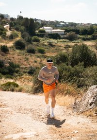 Atletický muž běží po hlinité cestě, má na sobě šedé tričko s dlouhým rukávem a oranžové kraťasy, černou čepici a bílé tenisky. V pozadí je zelené vegetace.