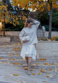 Bež džemper oversized kroja, bijela asimetrična suknja s čipkastim rubom, bež čizme do koljena, drži bijelu torbicu, raspršeni jesenski listovi.