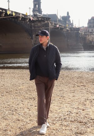 Hombre caminando por una orilla de guijarros cerca de un río, vistiendo una chaqueta de napa azul marino, suéter negro, pantalones marrones y zapatillas blancas; luz suave del sol.