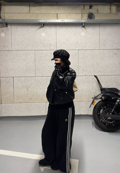 Chaqueta bomber de cuero negro, gorra negra, pantalones deportivos negros oversize con rayas blancas y calzado negro. Pared de concreto gris de fondo.