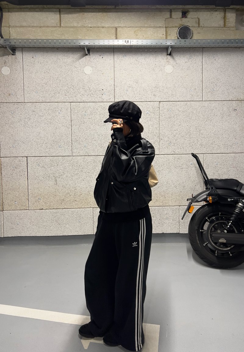 Veste aviateur en cuir noir, casquette noire, pantalon de survêtement noir oversize avec des rayures blanches, et chaussures noires. Mur en béton gris en arrière-plan.