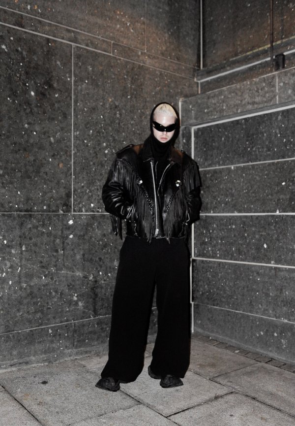Black leather jacket with fringe details, paired with a black turtleneck and wide-leg black pants. Accessories include dark sunglasses.