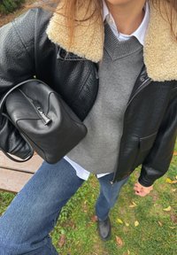 Crna kožna jakna s krem ovratnikom od ovčje kože preko sive veste, uparen s plavim trapericama i crnom torbom s teksturom.