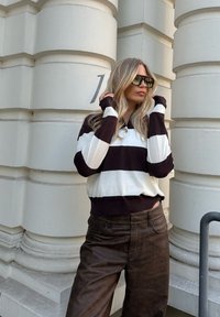 Gestreifter Pullover in Creme und Dunkelbraun mit Kragen und Bündchen. Braune Lederhosen. Stehend an einer säulenverzierten Wand.