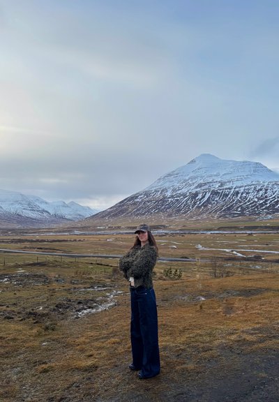 Suéter de punto en verde oscuro, jeans de pierna ancha oscuros y una gorra de camuflaje. Montañas cubiertas de nieve y terreno cubierto de hierba en el fondo con cielo nublado.