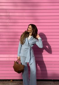 Abito azzurro con dettaglio con cintura annodata in vita, maniche lunghe; borsa marrone; sfondo con parete in legno rosa; ombre proiettate sul pavimento.