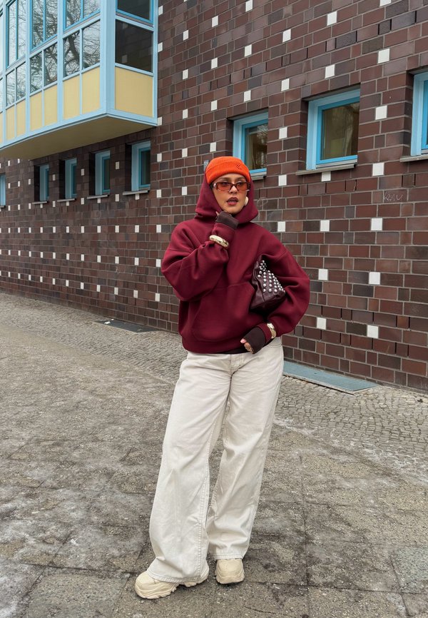 Person wearing orange beanie, burgundy hoodie, white wide pants, beige shoes, and sunglasses, standing against a brick wall with blue windows.