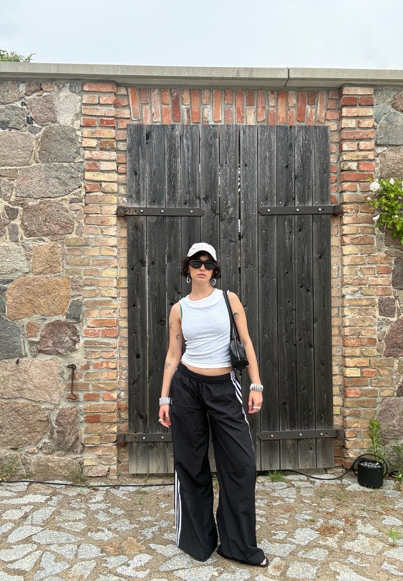 Personne portant un haut blanc sans manches, un pantalon noir à jambes larges, une casquette blanche et des lunettes de soleil, se tient devant de grandes portes en bois sombre encadrées par un mur de briques et de pierres.