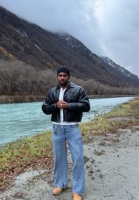 Veste courte en cuir noir portée sur une chemise blanche, associée à un jean large bleu clair et des bottes camel ; debout dans un paysage naturel près de l'eau.