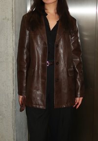 Femme portant un blazer en cuir marron foncé sur un haut et un pantalon noirs, debout devant un mur en béton et une porte en métal.