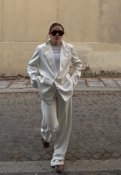 Mujer caminando por una calle empedrada con un traje blanco holgado, top blanco, grandes gafas de sol oscuras y zapatos marrones.