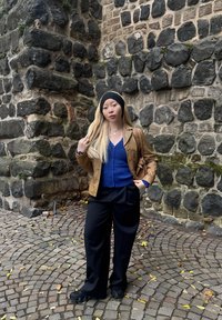 Brązowa skórzana kurtka, niebieska koszula z guzikami, czarne szerokie spodnie i czarna opaska na głowę. Stoi pod murami kamiennymi na kostce brukowej.