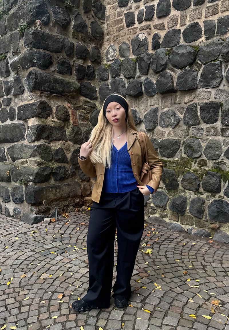 Hnědá kožená bunda, modrá košile na knoflíky, černé široké kalhoty a černý čelenka. Stojící proti kamenné zdi na kostkách.