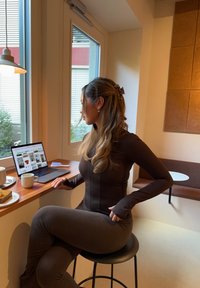 Brązowy sportowy top z wysokim kołnierzem i mankietami z otworem na kciuk, w zestawie z dopasowanymi legginsami. Siedzi na czarnym stołku obok laptopa i jedzenia.