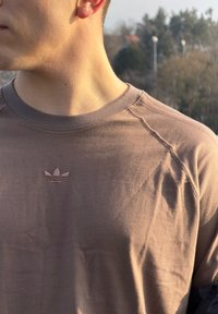 Barna pamut póló kerek nyakkivágással és látható varrási részletekkel, a mellkason tónusban hímzett Adidas logóval.