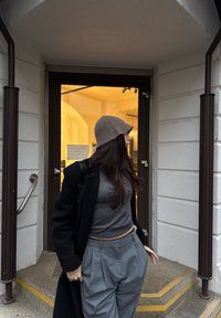 Personne portant un haut court gris et un pantalon plissé gris, coiffée d'un chapeau marron clair, debout près d'une porte vitrée à cadre marron avec une lumière jaune.