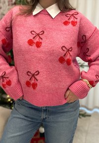 Vaaleanpunainen neulottu villapaita, jossa on punainen kirsikkakuosi ja ruskeat rusetit. Paidassa on resorit helman ja hihansuiden kohdalla, ja se on kerrostettu valkoisen kauluspaidan päälle.