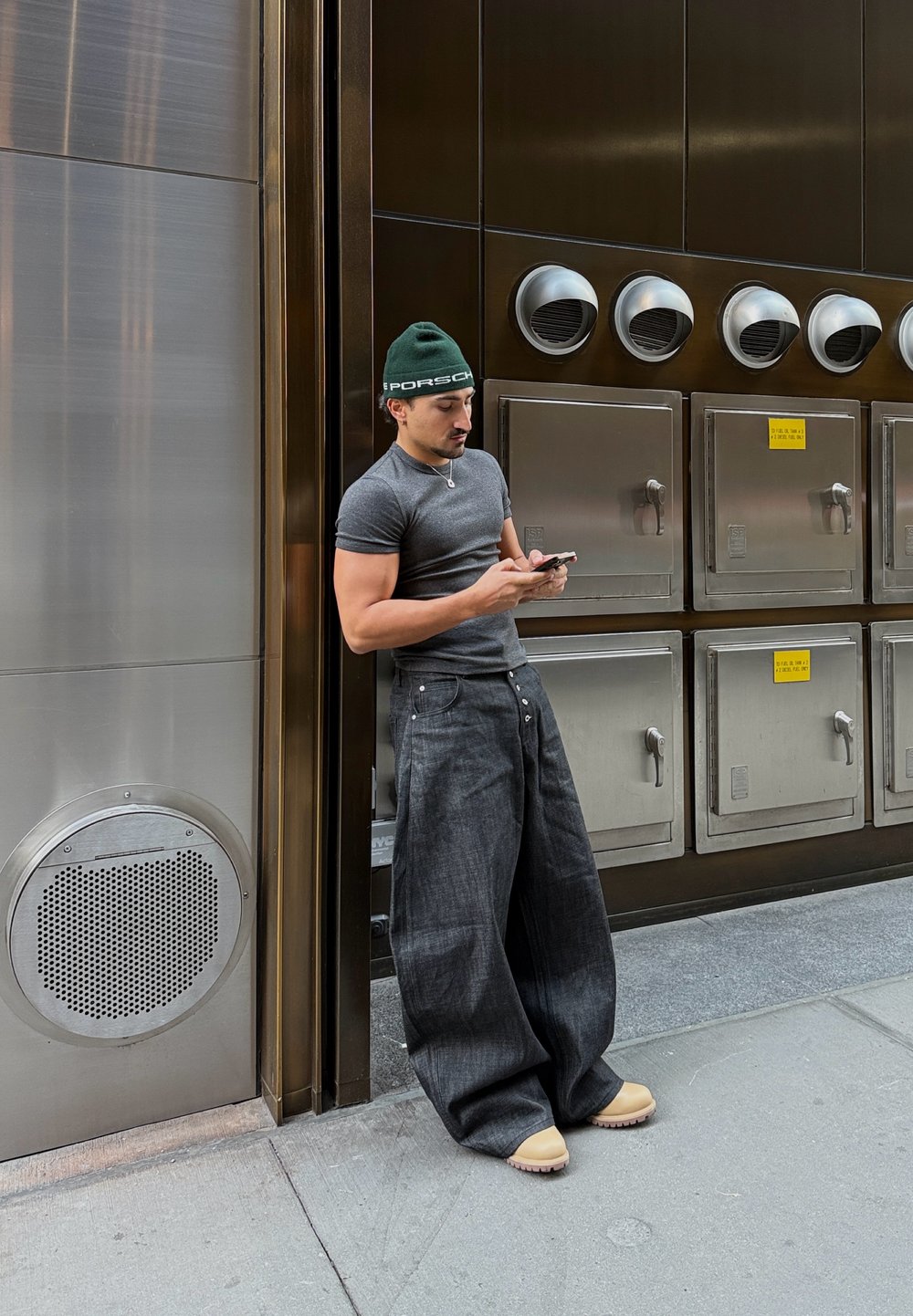 Muž v sivej priliehavej košeli a vo voľných tmavých džínsach, s zelenou čiapkou, sa opiera o kovovú stenu a používa smartfón.